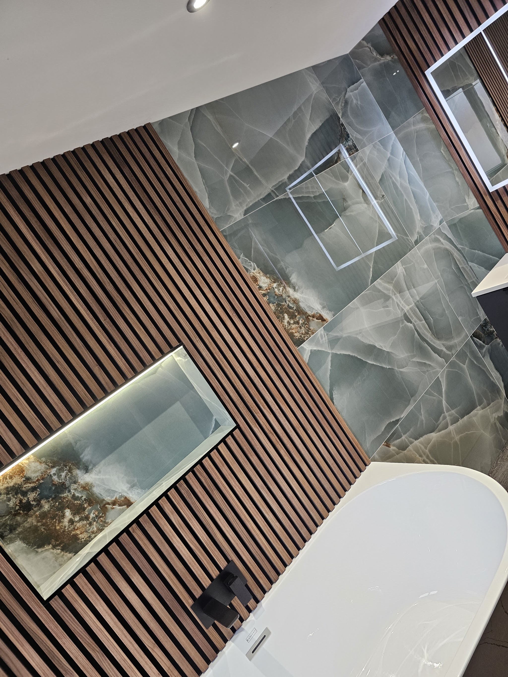 Freestanding bath beside a walnut slat wall panel with a lit niche and onyx-effect tiles